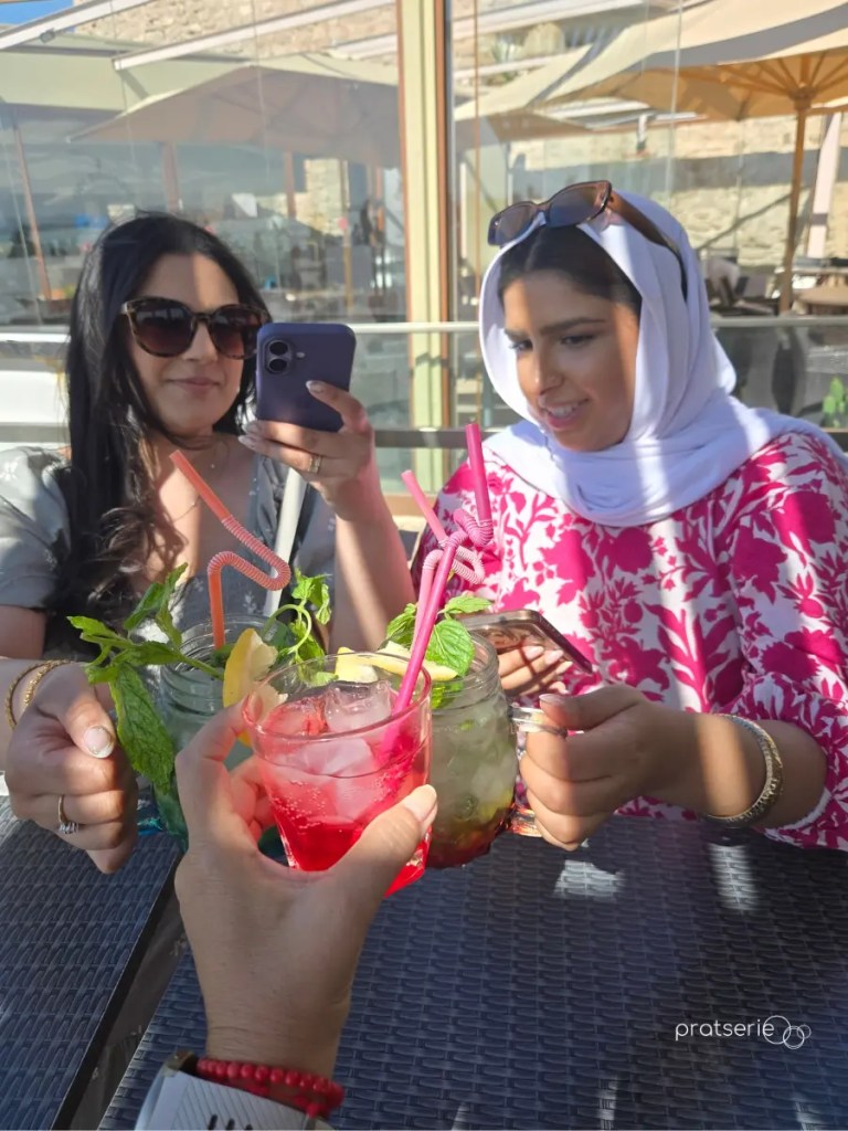 Três garotas fazendo um brinde com copos de bebidas coloridas com hortelã em uma mesa de café ao ar livre.