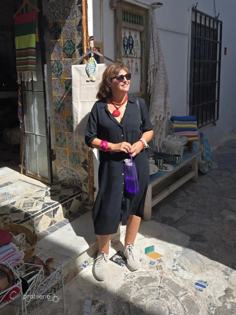 Mulher de vestido preto, óculos escuros e colar vermelho em pé sob o sol na entrada de uma loja decorada com azulejos tunisianos.